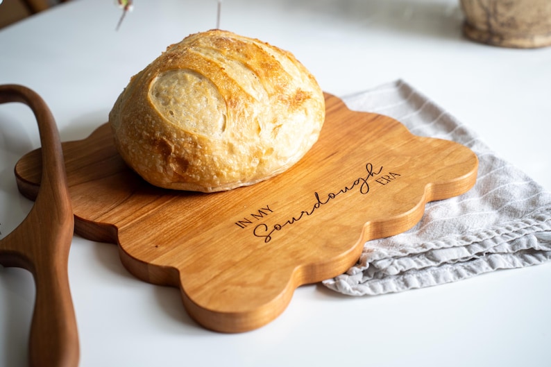 May include: A wooden cutting board with a scalloped edge, engraved with the text "In My Sourdough Era". A loaf of sourdough bread sits on the board.