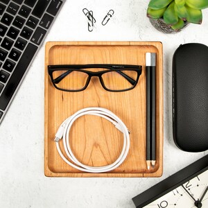 May include: A wooden tray with a white charging cable, black glasses, and two black pencils. The tray is on a white surface with a keyboard, a black glasses case, and a clock.