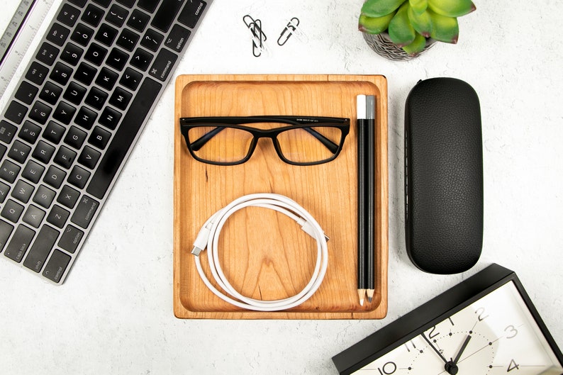 May include: A wooden tray with a white charging cable, black glasses, and two black pencils. The tray is on a white surface with a keyboard, a black glasses case, and a clock.