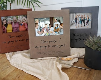 May include: Three photo albums in brown, tan, and gray, each with a framed photo and the text "These people are going to miss you!" The albums are on a light-colored surface with a plant and silverware.