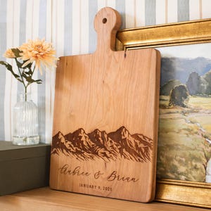 May include: A wooden cutting board with a mountain range design and the text "Aubree & Brian January 9, 2025" engraved on it.