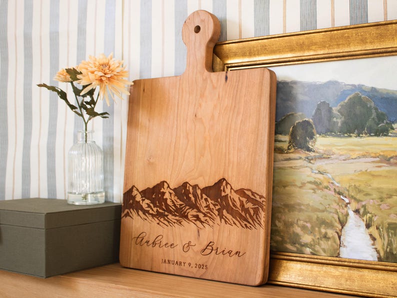 May include: A wooden cutting board with a mountain range design and the text "Aubree & Brian January 9, 2025" engraved on it.