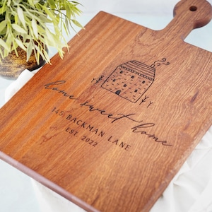 May include: A wooden cutting board with a house design and the text "home sweet home" and "145 Backman Lane Est. 2022" engraved on it.