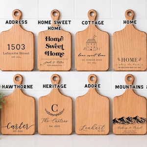 May include: Eight light brown wooden cutting boards with handles, arranged in two rows. Each board features unique black laser-engraved text and graphics, including addresses, names, dates, and illustrations like a house and mountain range.