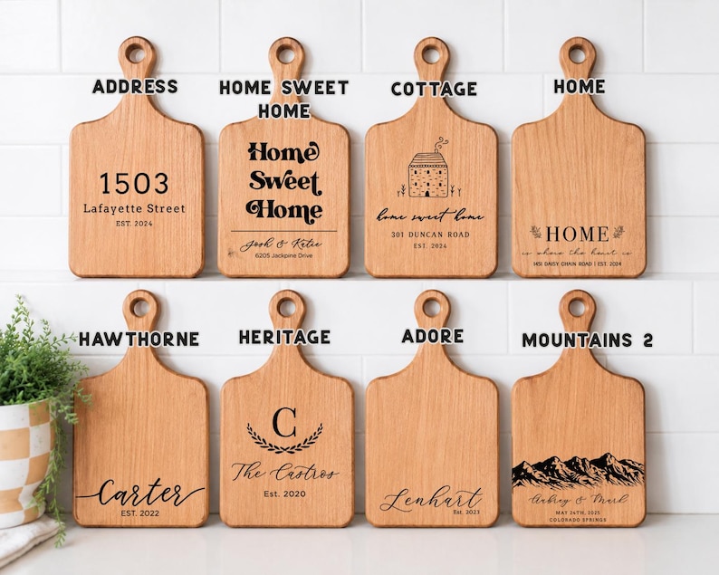 May include: Eight light brown wooden cutting boards with handles, arranged in two rows. Each board features unique black laser-engraved text and graphics, including addresses, names, dates, and illustrations like a house and mountain range.