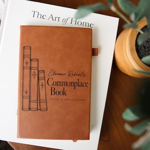May include: A tan leather-bound notebook featuring a black graphic of three stacked books and the text "Eleanor Roberts Commonplace Book NOTES & REFLECTIONS" printed on the front cover, with a small fabric pen loop attached to the side.