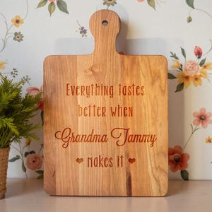 May include: A rectangular wooden cutting board with a rounded handle and a hole for hanging. The board is engraved with the text Everything tastes better when Grandma Tammy makes it, flanked by two small heart symbols on the bottom line.
