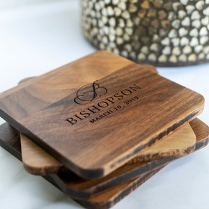 May include: Set of four square wooden coasters with a dark brown finish. The top coaster is engraved with the text "Bishopson March 10, 2019" and a decorative script letter "B".