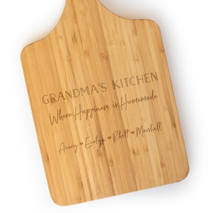 May include: A light brown bamboo cutting board with a handle, featuring the laser-engraved text "GRANDMA'S KITCHEN", "Where Happiness is Homemade", and the names "Avery", "Evelyn", "Rhett", and "Marshall" separated by small heart symbols.