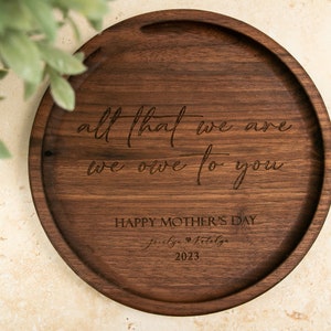 May include: A round wooden tray with the engraved message "all that we are we owe to you" and "Happy Mother's Day" with the date 2023.