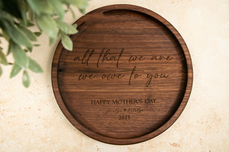 May include: A round wooden tray with the engraved message "all that we are we owe to you" and "Happy Mother's Day" with the date 2023.