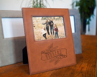 May include: A brown leather photo album with a family photo inside. The album has a rectangular frame and a banner with the name "RODGERS" engraved on it. A gray photo frame is visible in the background. The album is standing on a wooden surface.