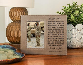 May include: A framed photo with a light brown border. The photo shows two people in military uniforms saluting. Text on the frame reads "I solemnly swear..." and "Enlisted June 16, 2023". The frame is on a wooden surface.