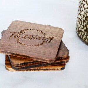 May include: Set of four wooden coasters with a dark brown finish. The top coaster features a laser-engraved design of a wreath with the word "Thesing" inside.