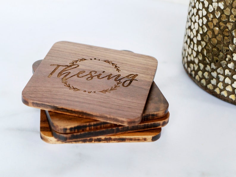 May include: Set of four wooden coasters with a dark brown finish. The top coaster features a laser-engraved design of a wreath with the word "Thesing" inside.