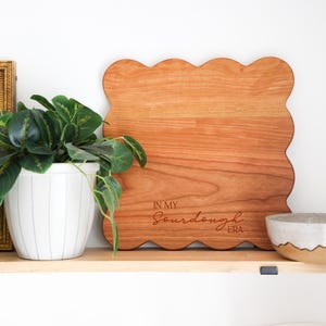 May include: A wooden cutting board with a scalloped edge and the text "In My Sourdough Era" engraved on the surface. The board is sitting on a shelf with a potted plant and a small bowl.
