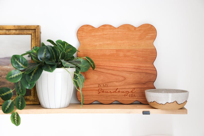 May include: A wooden cutting board with a scalloped edge and the text "In My Sourdough Era" engraved on the surface. The board is sitting on a shelf with a potted plant and a small bowl.
