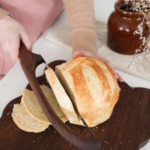 May include: A close-up of a loaf of sourdough bread being sliced on a dark brown wooden cutting board with a bread knife. The bread is light brown and has a crusty exterior.
