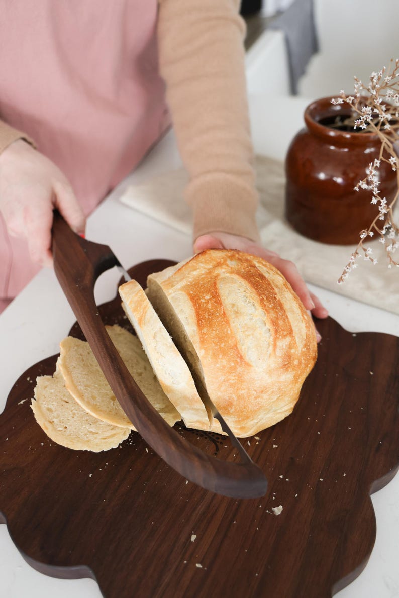 May include: A close-up of a loaf of sourdough bread being sliced on a dark brown wooden cutting board with a bread knife. The bread is light brown and has a crusty exterior.