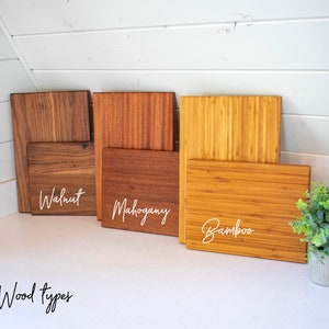 May include: A display of wooden cutting boards in walnut, mahogany, and bamboo. Each board is labeled with its wood type in white script. The boards are arranged on a white surface, with a small potted plant in the corner. Text at the bottom reads "Our Wood types."