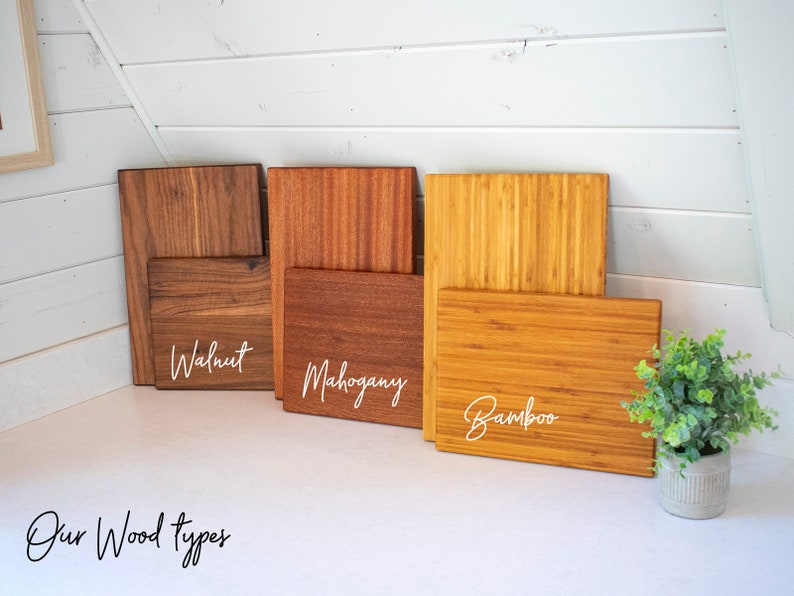 May include: A display of wooden cutting boards in walnut, mahogany, and bamboo. Each board is labeled with its wood type in white script. The boards are arranged on a white surface, with a small potted plant in the corner. Text at the bottom reads "Our Wood types."
