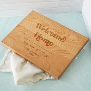 May include: A rectangular light brown wooden cutting board featuring the engraved text Welcome Home, followed by the names Luanne & Lucy, the address 1096 Greendale Ln., and the year 2022, all displayed in a mix of bold and script fonts.