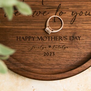 May include: A wooden ring dish with the text "To the one we love to you" and "Happy Mother's Day" engraved on it. The dish also has the names "Jocelyn & Katelyn" and the year "2023" engraved on it. A gold ring with a clear stone is sitting on the dish.