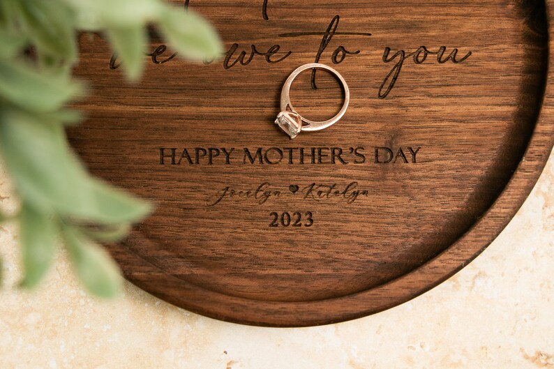 May include: A wooden ring dish with the text "To the one we love to you" and "Happy Mother's Day" engraved on it. The dish also has the names "Jocelyn & Katelyn" and the year "2023" engraved on it. A gold ring with a clear stone is sitting on the dish.