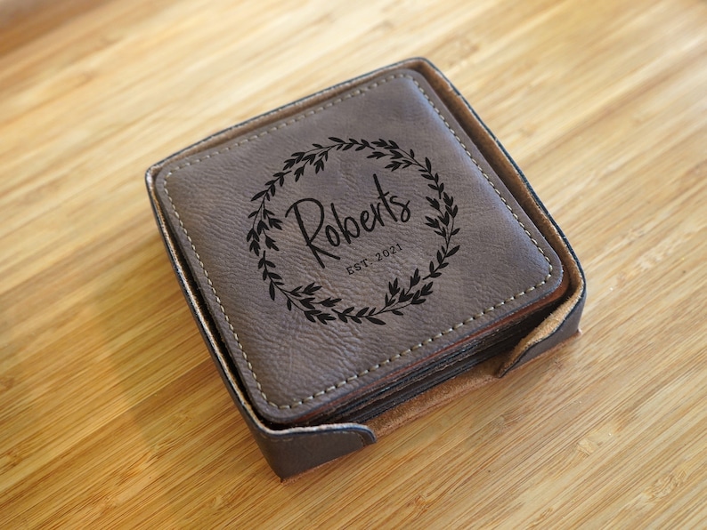 May include: A set of square, brown leather coasters with a holder. Each coaster features a black floral wreath design with the name "Roberts" and the year "EST. 2021". The coasters are in a matching brown holder.