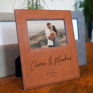 May include: A brown leather photo frame with a picture of a couple embracing. The frame is personalized with the names "Carrie & Markus" and the date "12 August 2017". A gray frame is visible in the background.