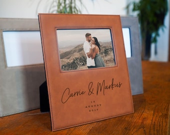 May include: A brown leather photo frame with a picture of a couple embracing. The frame is personalized with the names "Carrie & Markus" and the date "12 August 2017". A gray frame is visible in the background.