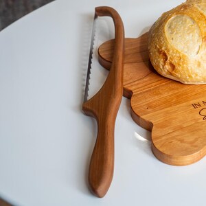 May include: A wooden bread knife with a serrated blade and a wooden handle. The knife is on a wooden cutting board with a loaf of bread on top. The cutting board has the words "In My Kitchen" engraved on it.