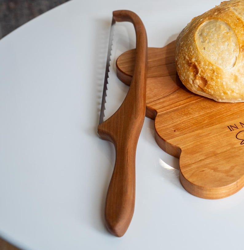 May include: A wooden bread knife with a serrated blade and a wooden handle. The knife is on a wooden cutting board with a loaf of bread on top. The cutting board has the words "In My Kitchen" engraved on it.