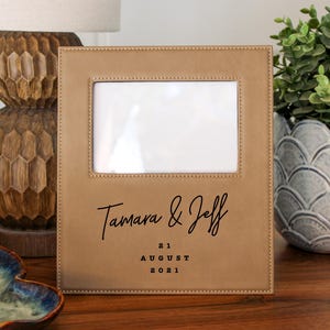 May include: A tan leather picture frame with a white photo area. The frame is personalized with the names "Tamara & Jeff" and the date "21 August 2021" in black script. The frame is on a wooden surface.