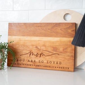 May include: A rectangular wooden cutting board with a warm brown grain, engraved with the word "mom" in script, the text "YOU ARE SO LOVED," and a list of names separated by heart symbols: Thor, Kate, Rory, Amy, Angelica, and Darick.