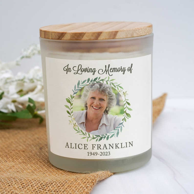 K&ouml;nnte beinhalten: Ein mattes Glas-Kerzengef&auml;&szlig; mit einem Holzdeckel. Das Gef&auml;&szlig; hat ein Etikett mit einem Foto einer Frau in einem Blumenkranz, dem Text "In Loving Memory of", "Alice Franklin" und den Daten "1949-2023".