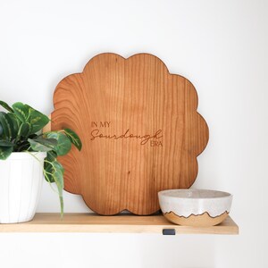May include: A wooden cutting board shaped like a flower with the text "In My Sourdough Era" engraved on it. The board is sitting on a wooden shelf with a white ceramic bowl and a green plant in a white pot.