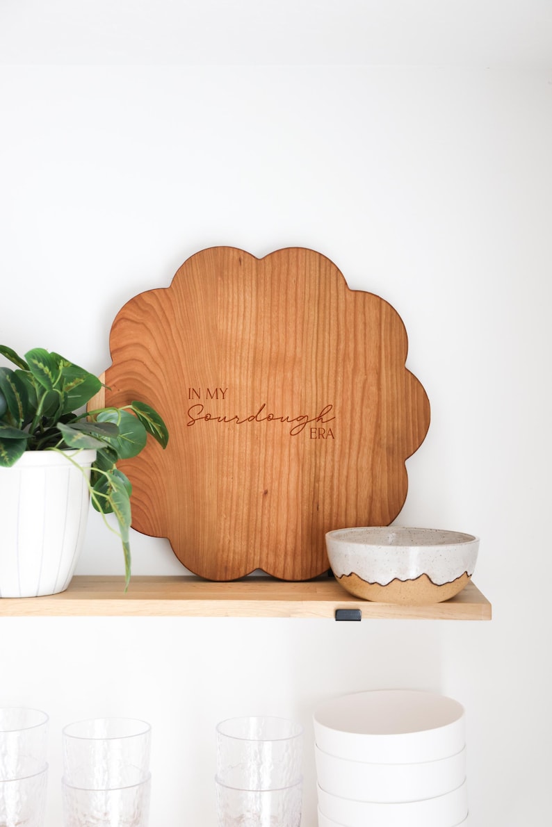 May include: A wooden cutting board shaped like a flower with the text "In My Sourdough Era" engraved on it. The board is sitting on a wooden shelf with a white ceramic bowl and a green plant in a white pot.