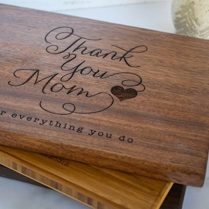 May include: A rectangular dark brown wooden cutting board with the engraved text Thank You Mom and a heart symbol, followed by the text for everything you do. The board is stacked on top of two other wooden boards of different wood tones.