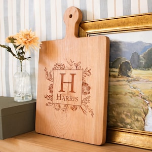 May include: A light-colored wooden cutting board with a handle and a hole for hanging. The board features a laser-engraved design of a square frame surrounded by floral illustrations, with the letter H and the name HARRIS centered inside.