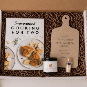 a gift box containing a cookbook, a wooden cutting board, a candle, and a small jar of spices.