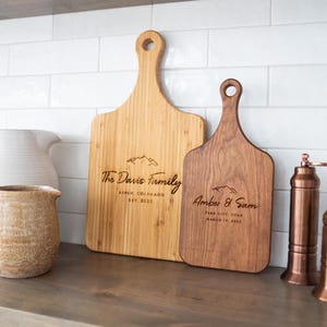 May include: Two personalized wooden cutting boards with engraved text. The larger board is light brown and says "The Davis Family, Aspen Colorado, Est 2023". The smaller board is dark brown and says "Amber & Sam, Park City Utah, Married in 2023".