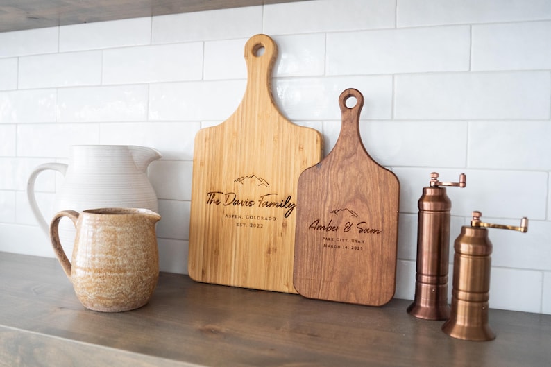 May include: Two personalized wooden cutting boards with engraved text. The larger board is light brown and says "The Davis Family, Aspen Colorado, Est 2023". The smaller board is dark brown and says "Amber & Sam, Park City Utah, Married in 2023".