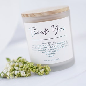 May include: A white frosted glass candle jar with a wooden lid. The label reads "Thank You" and includes a message of appreciation for a teacher. The candle is surrounded by small white flowers.