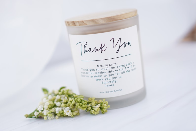 May include: A white frosted glass candle jar with a wooden lid. The label reads "Thank You" and includes a message of appreciation for a teacher. The candle is surrounded by small white flowers.