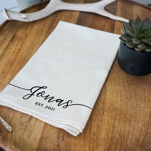 May include: Cream-colored kitchen towel with the name "Jonas" in elegant black script and the year "EST. 2021" below. The towel is folded and lies on a wooden surface, next to a small succulent in a black pot.