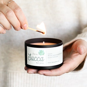 May include: A black candle with a white label that reads "I'm a Succa for you." The label also features a green succulent illustration and a short message. A person is lighting the candle with a match.
