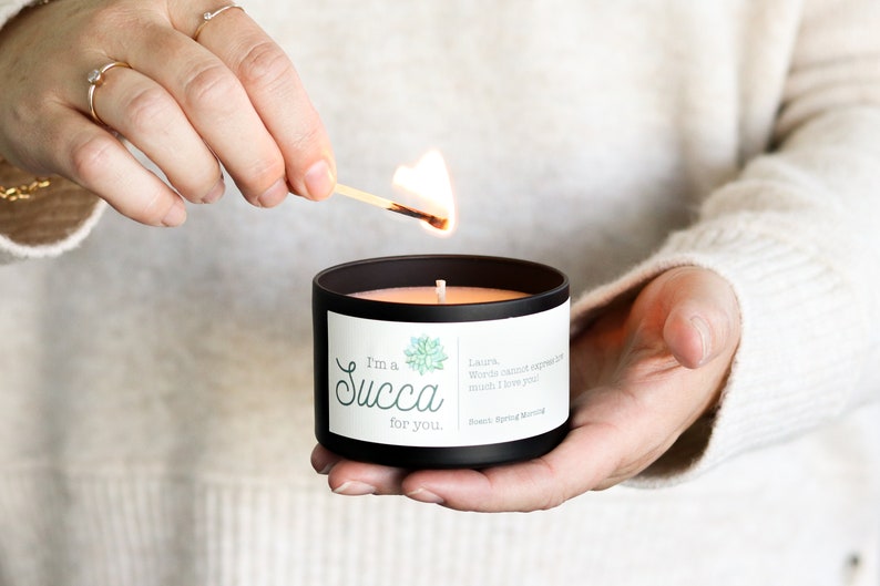 May include: A black candle with a white label that reads "I'm a Succa for you." The label also features a green succulent illustration and a short message. A person is lighting the candle with a match.