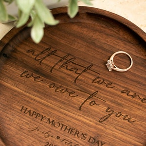 May include: A round wooden tray with the inscription "all that we are we owe to you" and "Happy Mother's Day" engraved on the surface. A gold ring with a clear stone is resting on the tray.