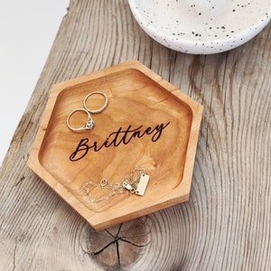 May include: A hexagonal wooden tray with the name "Brittney" engraved on it. The tray is filled with jewelry, including two rings and a necklace.